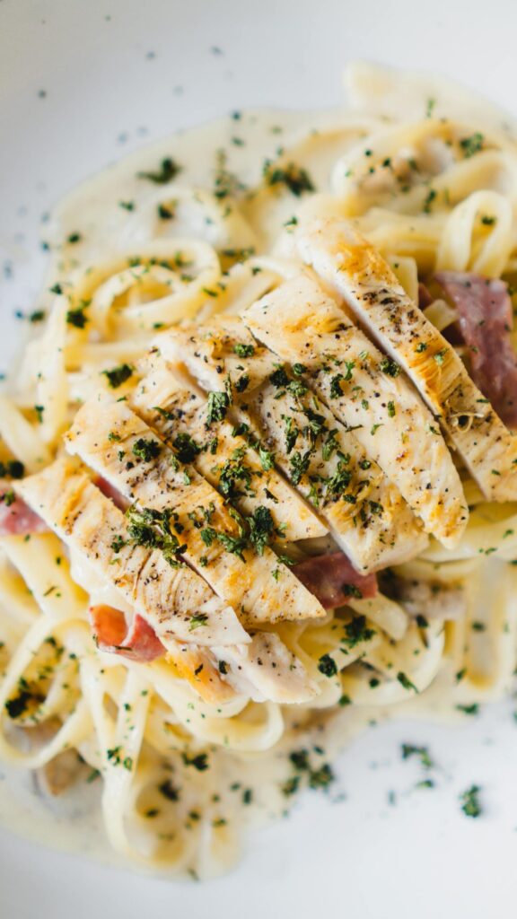 Close-up of a creamy pasta dish topped with grilled chicken slices, garnished with fresh parsley. The pasta is coated in a rich white sauce, with hints of ham and herbs visible beneath the chicken showing easy Italian dinners.