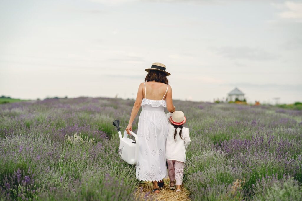 Image of a mother and child walking together in a lavender field, both wearing straw hats, with the mother carrying a white watering can. The scene is serene with a small house visible in the distance representing I feel like a bad mom because I yell.