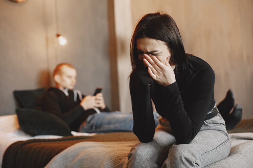 Image of a woman sits on the edge of a bed, covering her face with her hand, appearing distressed or upset. In the background, a man is sitting on the bed, focused on his phone, seemingly unaware of her emotions. The room has a warm, muted tone with soft lighting, highlighting the emotional distance between the two.