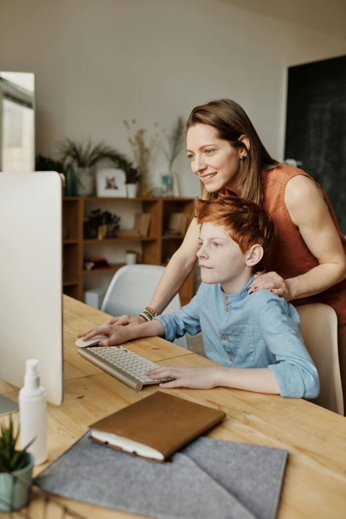 An adult woman and a young boy looking at a computer screen together, with the boy using the keyboard. They are sitting at a wooden desk in a cozy, well-lit room, with books and decorative items in the background.