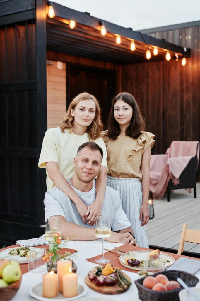 A family photo of three people at an outdoor dinner setting. A man is seated at the table with a relaxed smile, and a woman stands behind him with her arms gently on his shoulders. Next to her, a young girl stands with a calm expression. The table is set with candles, food, and wine glasses. String lights are hanging above, creating a warm, cozy atmosphere showing how do you express gratitude to your family.