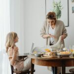 Close-up image featuring two women. One is seated with a laptop on her lap, gesturing animatedly as she talks. The other is standing, wearing a cozy cardigan, holding a dish towel, and appearing engaged in conversation. The table between them is set with bread, fruit, and breakfast items, while a botanical-themed framed artwork decorates the wall in the background representing Why Do I Feel Like a Bad Mom.