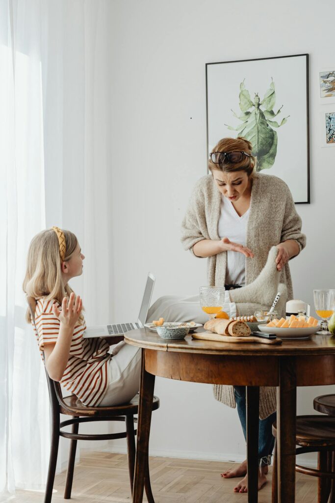 Close-up image featuring two women. One is seated with a laptop on her lap, gesturing animatedly as she talks. The other is standing, wearing a cozy cardigan, holding a dish towel, and appearing engaged in conversation. The table between them is set with bread, fruit, and breakfast items, while a botanical-themed framed artwork decorates the wall in the background representing Why Do I Feel Like a Bad Mom.