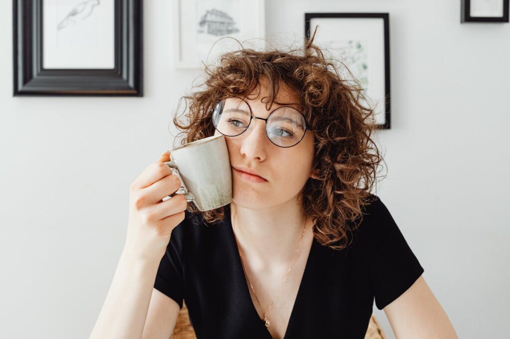 Image of a woman with curly hair wearing round glasses, holding a cup near their face. The person appears thoughtful, gazing into the distance. The background includes framed wall art in a minimalist setting showing feeling like a bad mom.