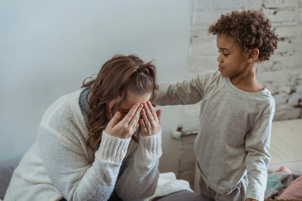 Image of a young child with curly hair gently comforting a woman in a sweater who has her face in her hands, set in a cozy indoor environment with a textured wall in the background.