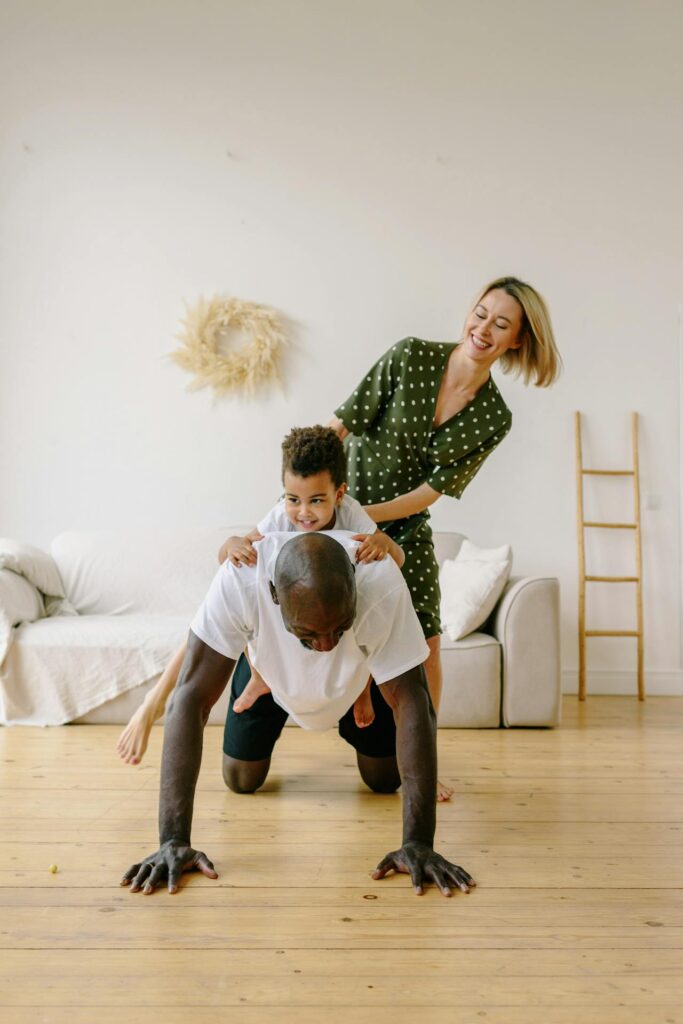 Image of a playful family scene in a living room with wooden floors. A man in a white T-shirt is in a push-up position while a child sits on his back, smiling. A woman in a green polka-dot dress stands beside them, laughing and holding the child for support. The background includes a sofa with a white throw, a decorative wreath on the wall, and a wooden ladder leaning against it.