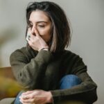 Image of a woman with short, dark hair sits with her knee up, wearing a dark green sweater and blue jeans. She appears to be lost in thought, covering part of her face with her hand. She has rings on her fingers, including a turquoise ring on her index finger. The background is simple and blurred, creating a soft, introspective mood. Image used for the article Why Do I Feel Disconnected From My Husband.