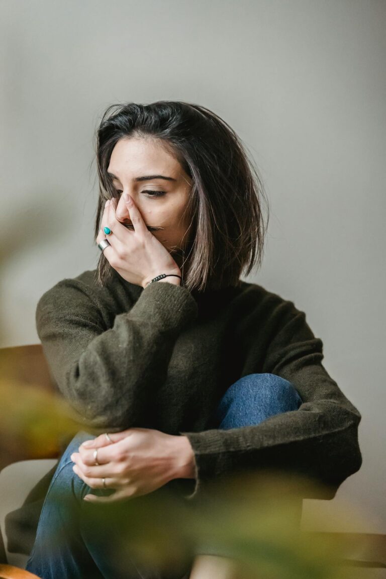 Image of a woman with short, dark hair sits with her knee up, wearing a dark green sweater and blue jeans. She appears to be lost in thought, covering part of her face with her hand. She has rings on her fingers, including a turquoise ring on her index finger. The background is simple and blurred, creating a soft, introspective mood. Image used for the article Why Do I Feel Disconnected From My Husband.