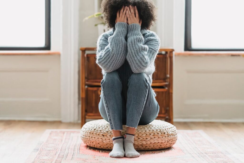 Image of a person sitting on a round woven cushion with knees pulled to their chest, covering their face with both hands. They are wearing a chunky gray sweater, jeans, and light-colored socks. The background shows a cozy room with natural light coming in from windows and a wooden cabinet in the corner.