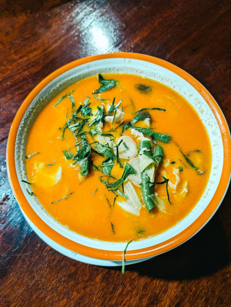 Image of a bowl of creamy orange soup, likely Thai Tom Kha Gai or a similar coconut milk-based soup. It contains pieces of chicken, green beans, and is garnished with thinly sliced fresh herbs, possibly kaffir lime leaves or cilantro. The dish is served in a white bowl with an orange rim, placed on a dark wooden table, creating a warm and appetizing presentation.