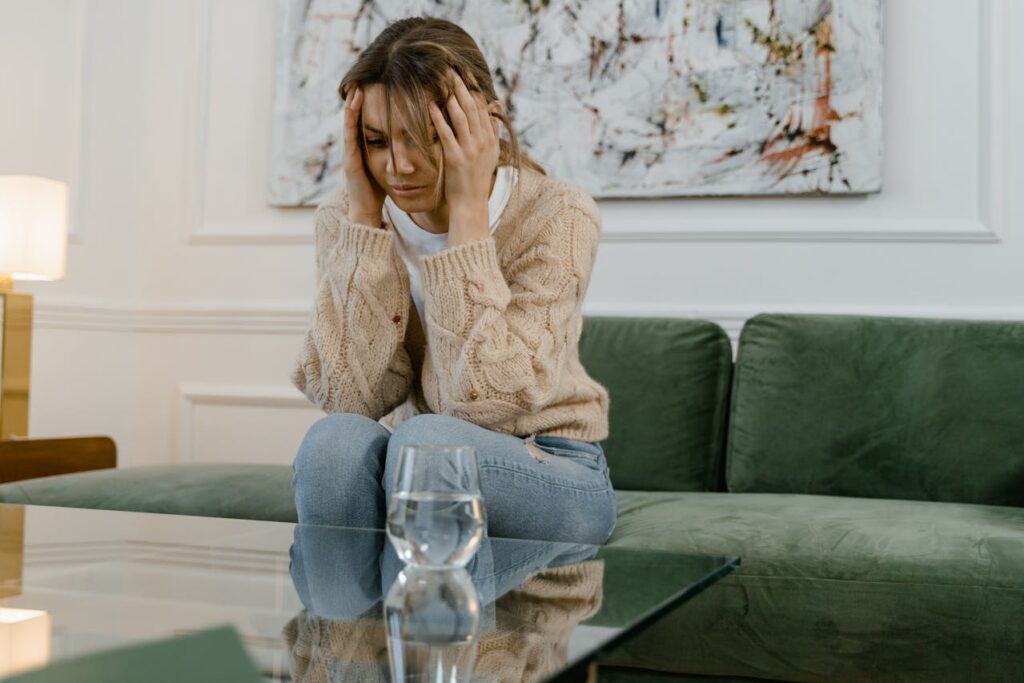 Image of a woman sitting on a green couch in a modern living room, holding her head with both hands, appearing distressed or deep in thought, with a glass of water on the glass table in front of her and abstract artwork on the wall.