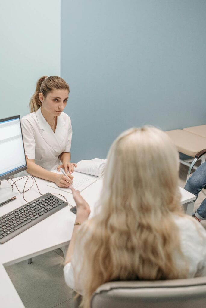 Image of two women in white dresses discussing a topic, there's a computer on the desk and one woman is also writing about the talk they both are having shows Postpartum depression and marriage.