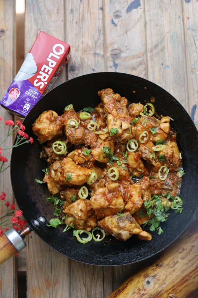 Image of serving chicken karahi, a popular South Asian dish, presented in a black wok. The dish features cooked chicken pieces in a rich, spiced tomato-based curry, garnished with fresh green chilies and chopped coriander. A carton of Olper's dairy cream is placed beside it on a rustic wooden surface, suggesting its use in the recipe. Bright flowers add a decorative touch to the setup.