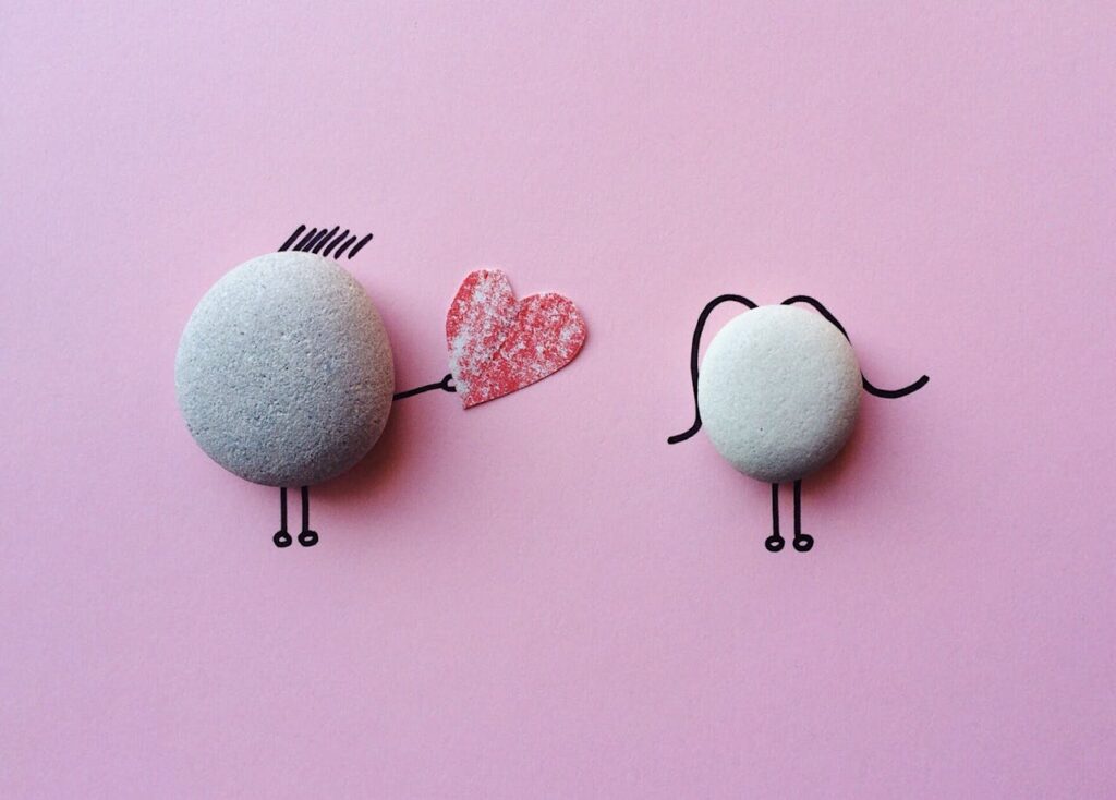 Image of two pebble figures on a pink background. The figure on the left, with small drawn lines for hair, is holding a red heart toward the figure on the right, which has curved lines suggesting long hair. Both figures have simple stick legs, giving the image a cute, playful feel.