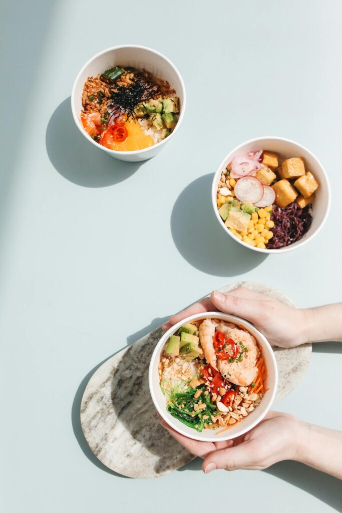 Image of three bowls of vibrant poke bowls are placed on a light pastel background. Each bowl is filled with fresh ingredients, including avocado, rice, vegetables, and toppings like seaweed and shrimp. A pair of hands is holding one bowl on a marble tray shows easy recipes for working moms.