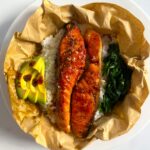 Image of a bowl of food containing glazed salmon fillets on a bed of white rice, accompanied by slices of fresh avocado and sautéed spinach. The dish is presented in parchment paper, placed on a white plate. Image used for the article 11 Easy Recipes For Busy Moms.