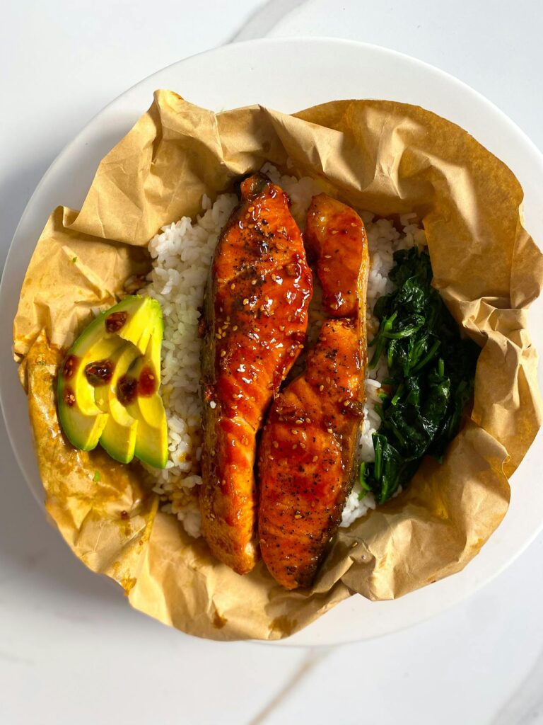 Image of a bowl of food containing glazed salmon fillets on a bed of white rice, accompanied by slices of fresh avocado and sautéed spinach. The dish is presented in parchment paper, placed on a white plate. Image used for the article 11 Easy Recipes For Busy Moms.