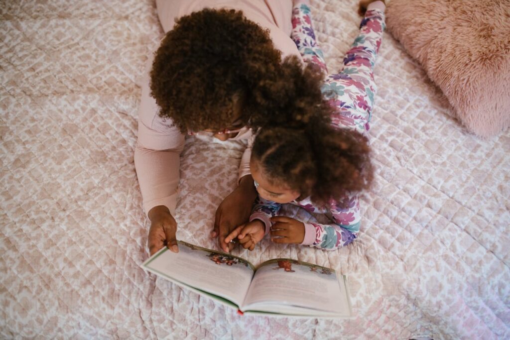 Image of an adult and a young child lying on a bed, reading a book together. The adult is wearing a light pink top, and the child is dressed in floral pajamas. They are both focused on the book, with the child's hand resting on the page. The bed has a quilted cover and a soft, fluffy pillow nearby.