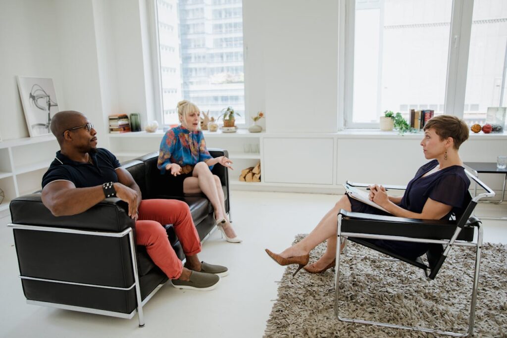 Image of couple sits on a couch during a therapy session. The woman, with blonde hair in a bun and wearing a blue floral top, appears to be speaking and gesturing with her hands. The man, wearing a black shirt and red pants, sits beside her, listening intently. Across from them sits a therapist, a woman with short brown hair, holding a notebook and pen, listening attentively. The room is bright and modern, with large windows and minimal decor, including shelves with a few books and decorative items.