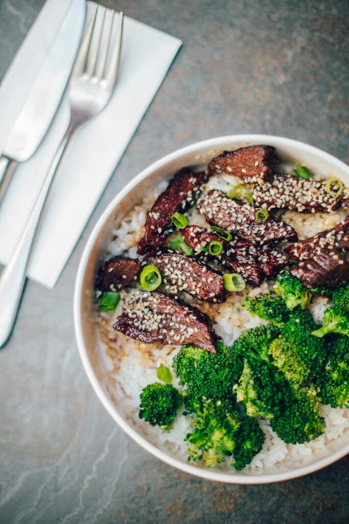 Close-up of a bowl of food featuring white rice topped with seared beef slices, garnished with sesame seeds and chopped green onions, alongside steamed broccoli. A fork and knife rest on a folded napkin beside the bowl representing Sunday evening dinner recipes.