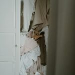 A partially open closet filled with a disorganized pile of clothes in neutral colors like beige, white, and tan. The clothes are crumpled and stacked haphazardly on a single shelf, creating a cluttered appearance. The closet door is white, and only part of it is visible. Image used for the article Can't Relax Until The House Is Clean.