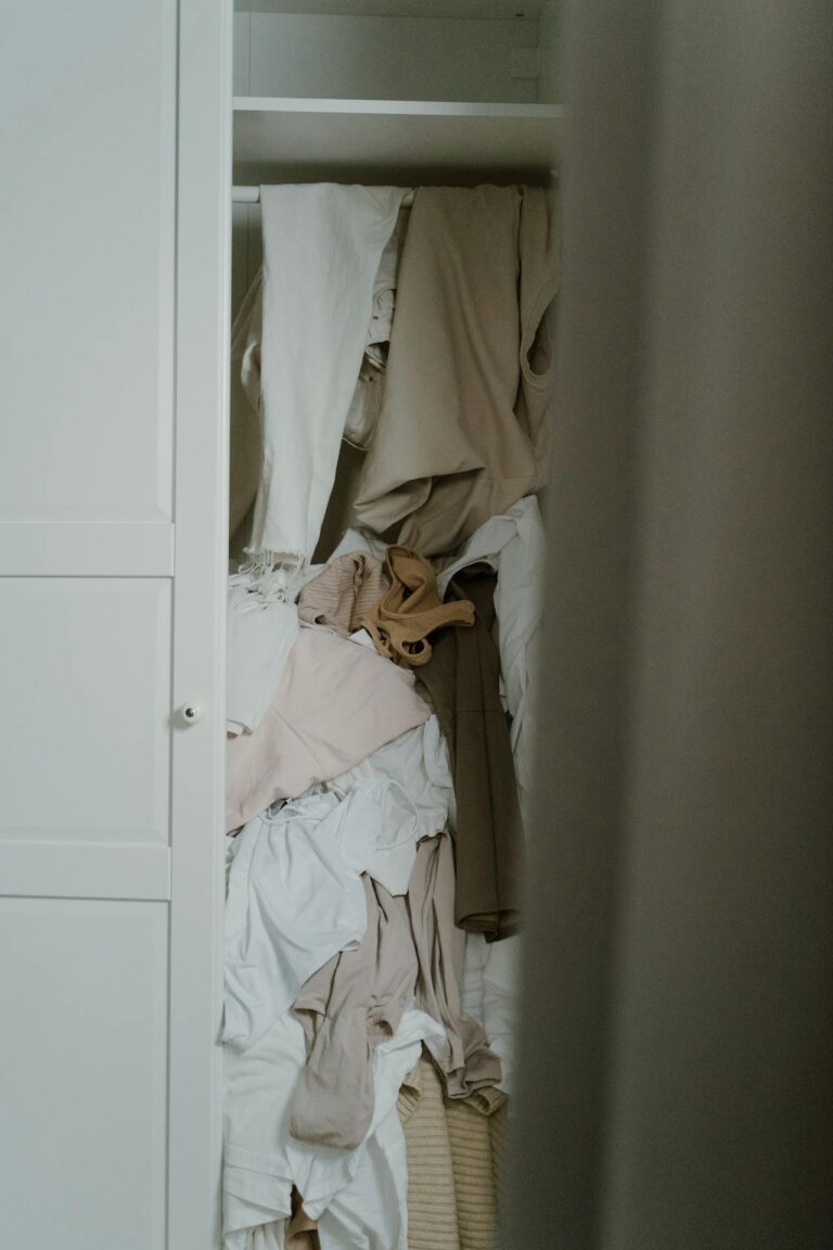 A partially open closet filled with a disorganized pile of clothes in neutral colors like beige, white, and tan. The clothes are crumpled and stacked haphazardly on a single shelf, creating a cluttered appearance. The closet door is white, and only part of it is visible. Image used for the article Can't Relax Until The House Is Clean.