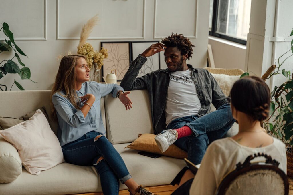 Image of a group of three people in a casual indoor setting, sitting on a beige couch and chair, engaged in a discussion. The room has a cozy and modern decor with plants, cushions, and soft lighting. One person gestures while speaking, another listens attentively, and a third person sits with their back to the camera.