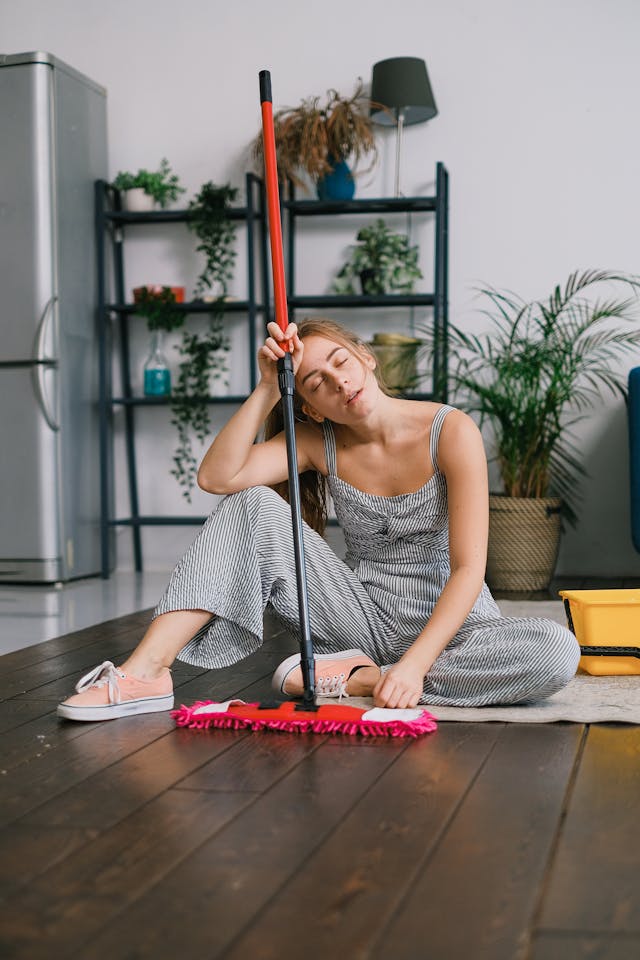 Image of a young woman sits on the floor, looking tired while holding a mop. She is dressed in a striped jumpsuit and sneakers, with her head resting on her hand and eyes closed. Behind her is a modern living space with plants, a black shelving unit with decorative items, and a gray refrigerator. A yellow cleaning bucket is on the floor next to her. The wooden floor is polished, and the room has a relaxed, homey feel showing decluttering checklist for the article can't relax until the house is clean.