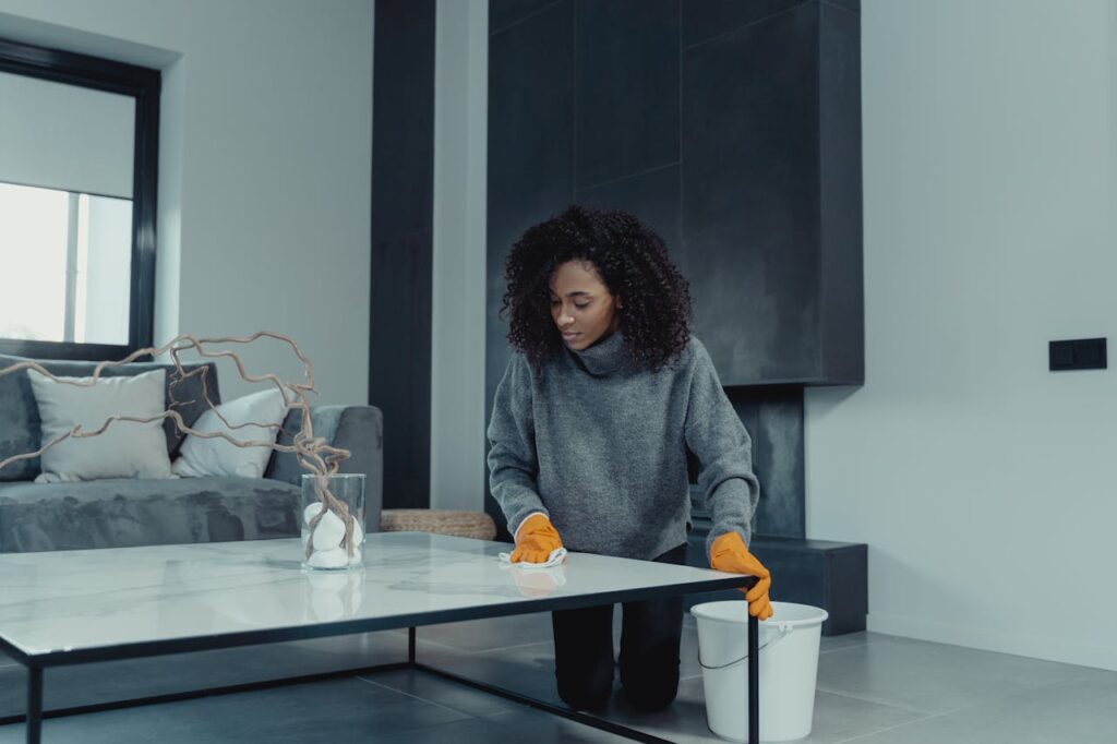 Image of a woman kneeling on the floor and cleaning a coffee table with a cloth. They are wearing a gray sweater and orange gloves, with a white bucket nearby. The room has a modern, minimalist design, featuring a gray sofa with pillows and a glass vase containing decorative branches on the table. Image used for article Why am I obsessed with cleaning my house.