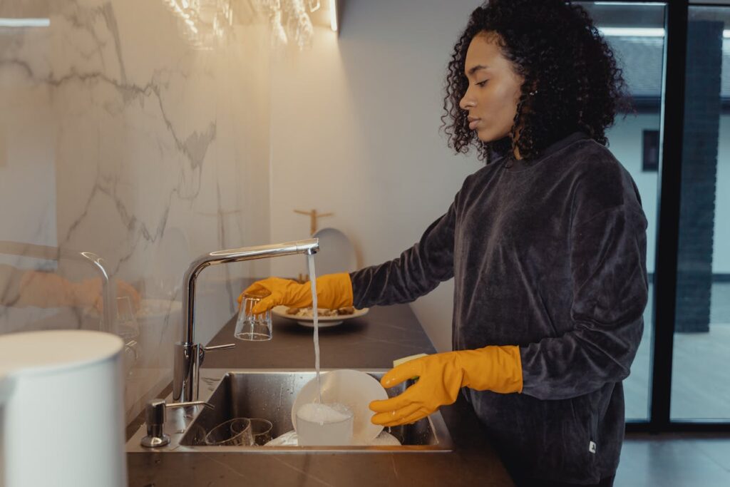 Image of a woman washing dishes in a modern kitchen while wearing yellow rubber gloves. She is focused on cleaning a glass under running water from the faucet. The kitchen features a sleek, marble-patterned backsplash and a minimalist design.