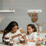 Image of a woman and a young girl decorating pastries together at a table in a cozy kitchen. The table is filled with ingredients, colorful eggs, and festive decorations, creating a warm and creative atmosphere representing Characteristics Of A Good Mom.