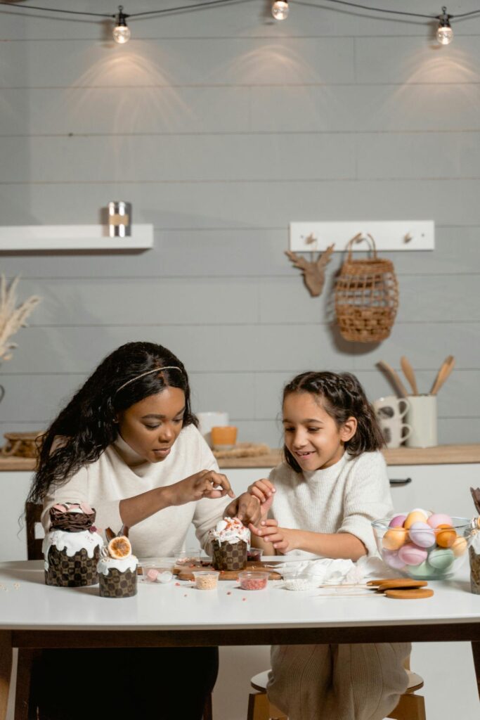 Image of a woman and a young girl decorating pastries together at a table in a cozy kitchen. The table is filled with ingredients, colorful eggs, and festive decorations, creating a warm and creative atmosphere representing Characteristics Of A Good Mom.