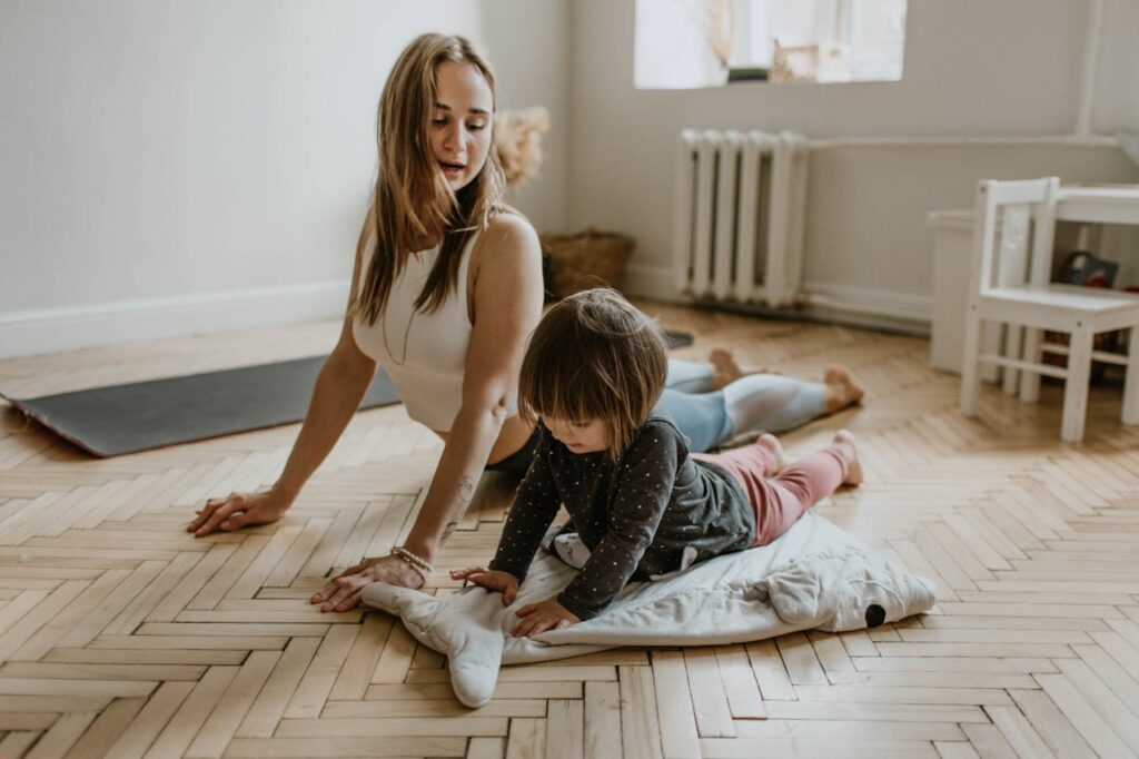 Close-up image of a woman and a young child practicing yoga together on a wooden floor in a bright room. The child is lying on a soft mat, mimicking a stretch, while the woman guides them with care and encouragement representing mom traits