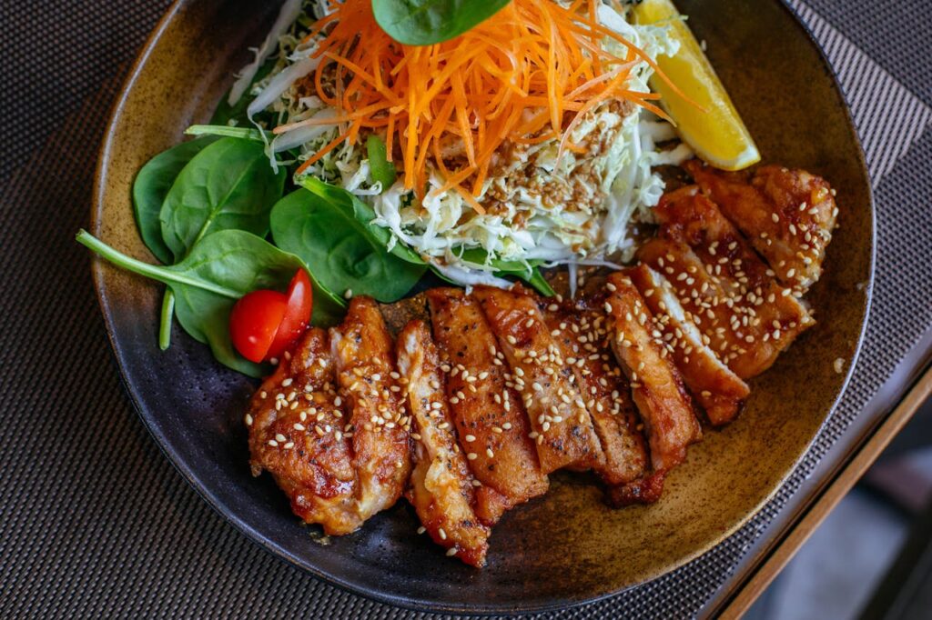 Image of a dish featuring sliced grilled chicken topped with sesame seeds, accompanied by a fresh salad of shredded cabbage, julienned carrots, spinach leaves, and cherry tomatoes. A wedge of lemon is placed on the side for garnish. The meal is served in a rustic, earthy-toned bowl.