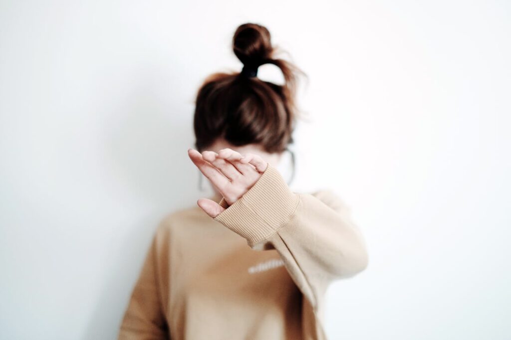 Image of a woman with their hair in a messy bun, wearing a beige sweater, raises their hand in front of their face as if blocking the camera. The background is plain white, emphasizing a minimalist and candid atmosphere representing  disappointed in husband after baby.