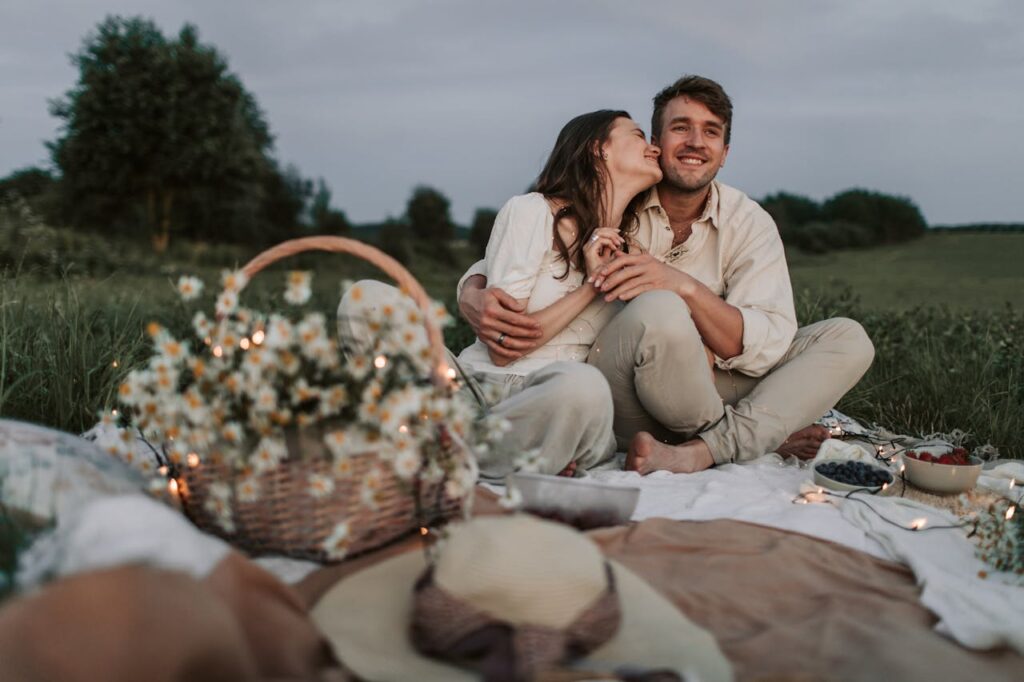 Image of a couple enjoys a romantic outdoor picnic on a blanket in a scenic, grassy field. The woman, smiling, leans in to kiss the man's cheek while he wraps an arm around her and smiles happily. In the foreground, there’s a woven basket filled with daisies, hats, and a spread of food, including berries and other small dishes. The atmosphere is serene, with soft evening light adding a warm and intimate touch.