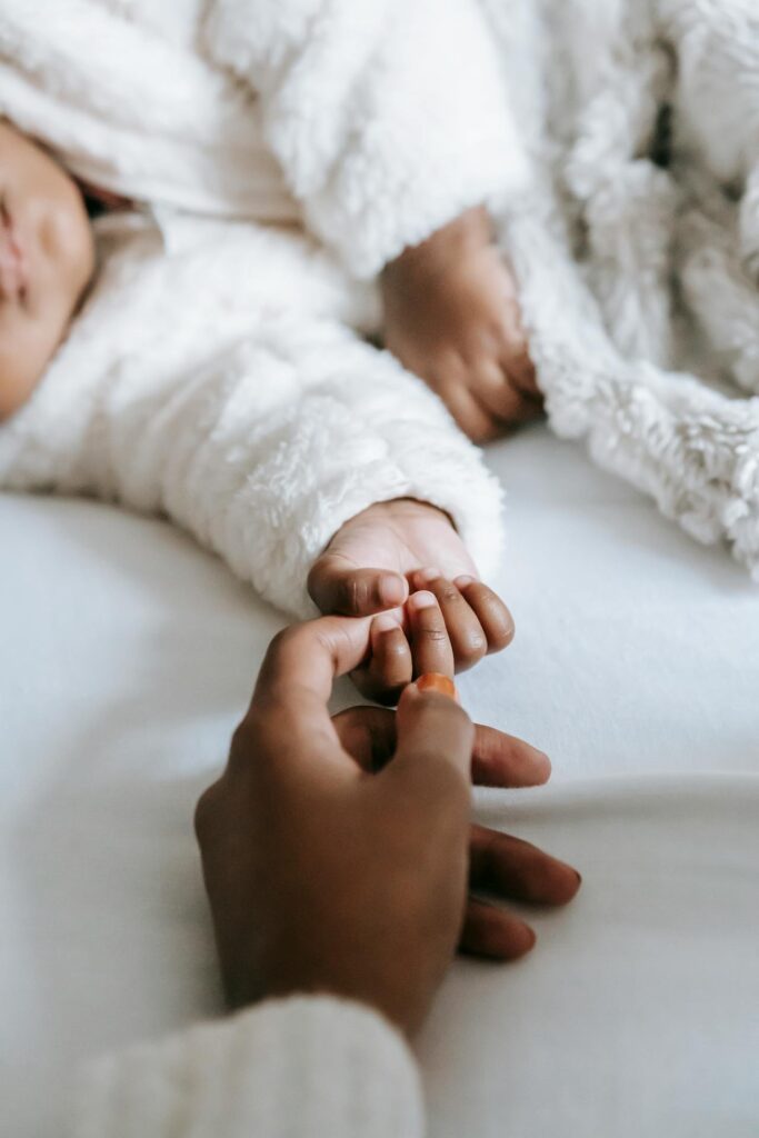 Image of a baby lying on a white bed, dressed in soft, fluffy white clothing. An adult gently holds the baby's tiny hand, creating a warm and intimate connection. The scene conveys comfort, love, and care showing  unsupportive husband postpartum.