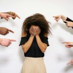 Image of a women standing with their face covered in their hands, surrounded by multiple hands pointing accusatory fingers at them, conveying a sense of blame or judgement Image used for the article Postpartum Resentment Towards Husband.