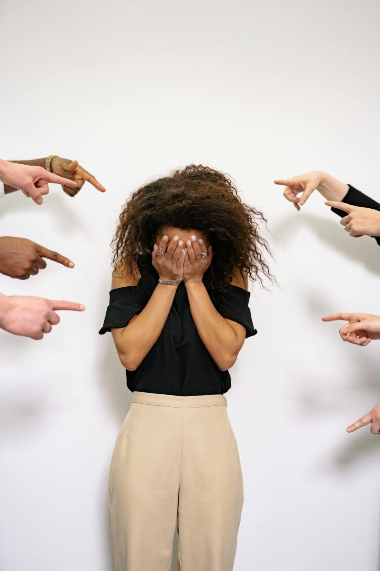 Image of a women standing with their face covered in their hands, surrounded by multiple hands pointing accusatory fingers at them, conveying a sense of blame or judgement Image used for the article Postpartum Resentment Towards Husband.