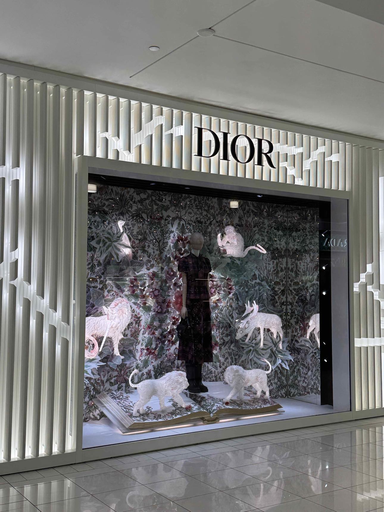 Image of a Dior store window display with a mannequin, white animal sculptures, an open book, and floral patterns.