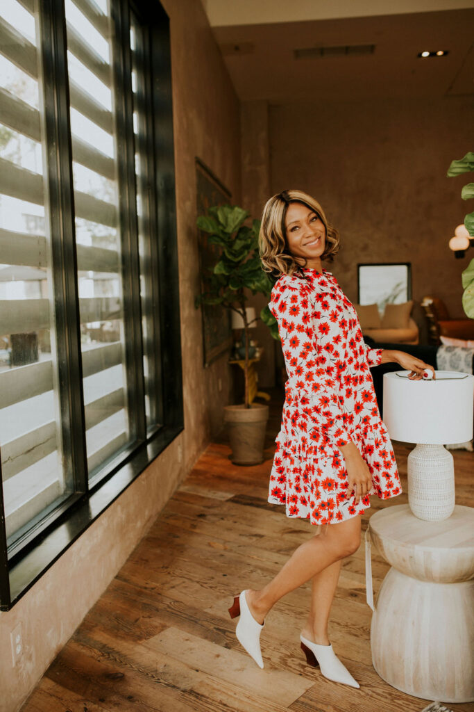 Image of a woman in a bright floral dress stands smiling in a modern room with wooden floors and large windows. Image used for the article A Day in the Life of a Mom.