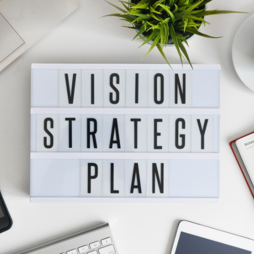 Image of black letters spelling, VISION STRATEGY PLAN, sits on a white desk. Surrounding the lightbox are various work essentials: a small green plant at the top, a partially visible laptop, a notebook with a red border, and other office items, creating a clean and organized workspace, showing Couple Vision Board.