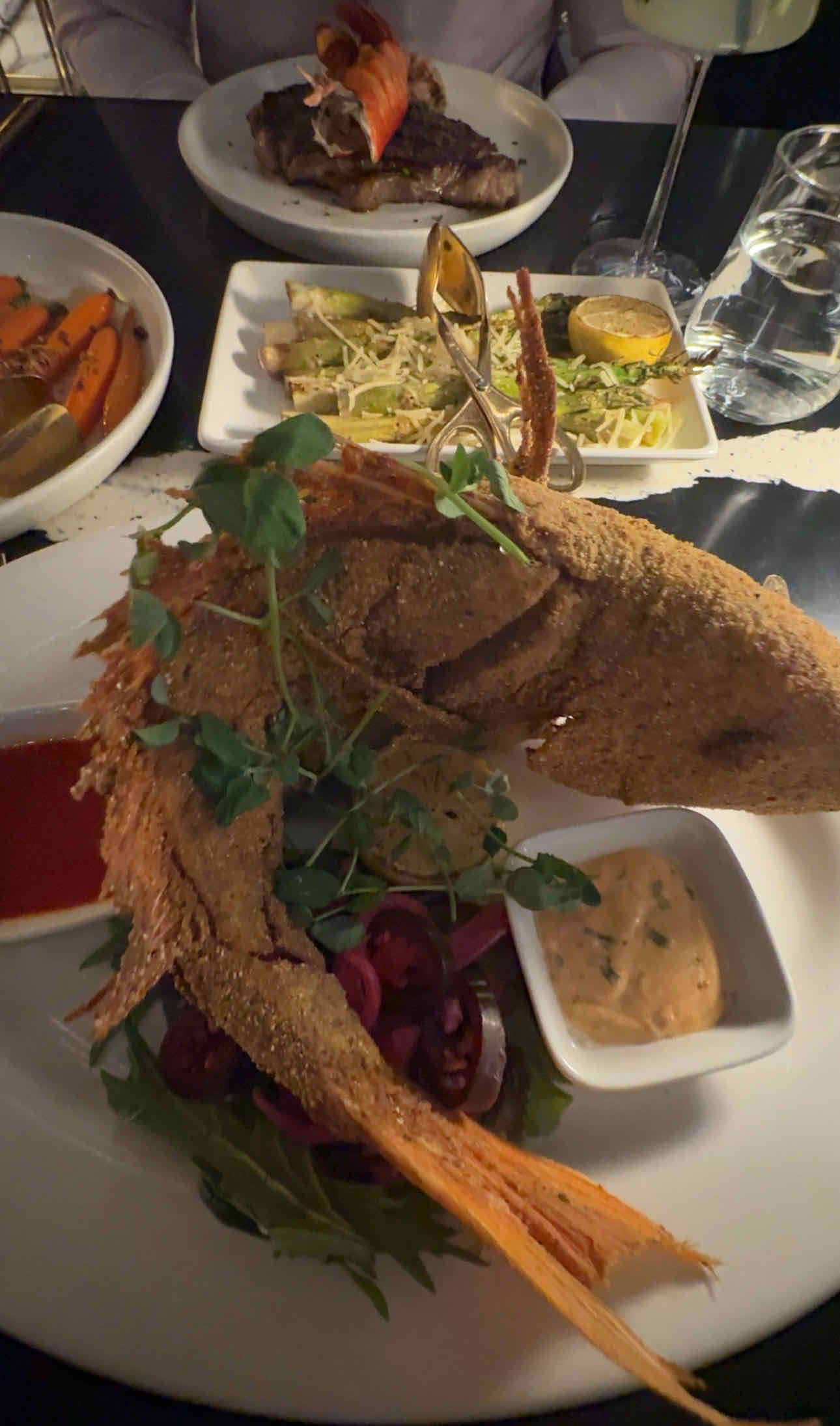 Image of a fried fish served with sauces, greens, and a side of lobster and grilled vegetables.