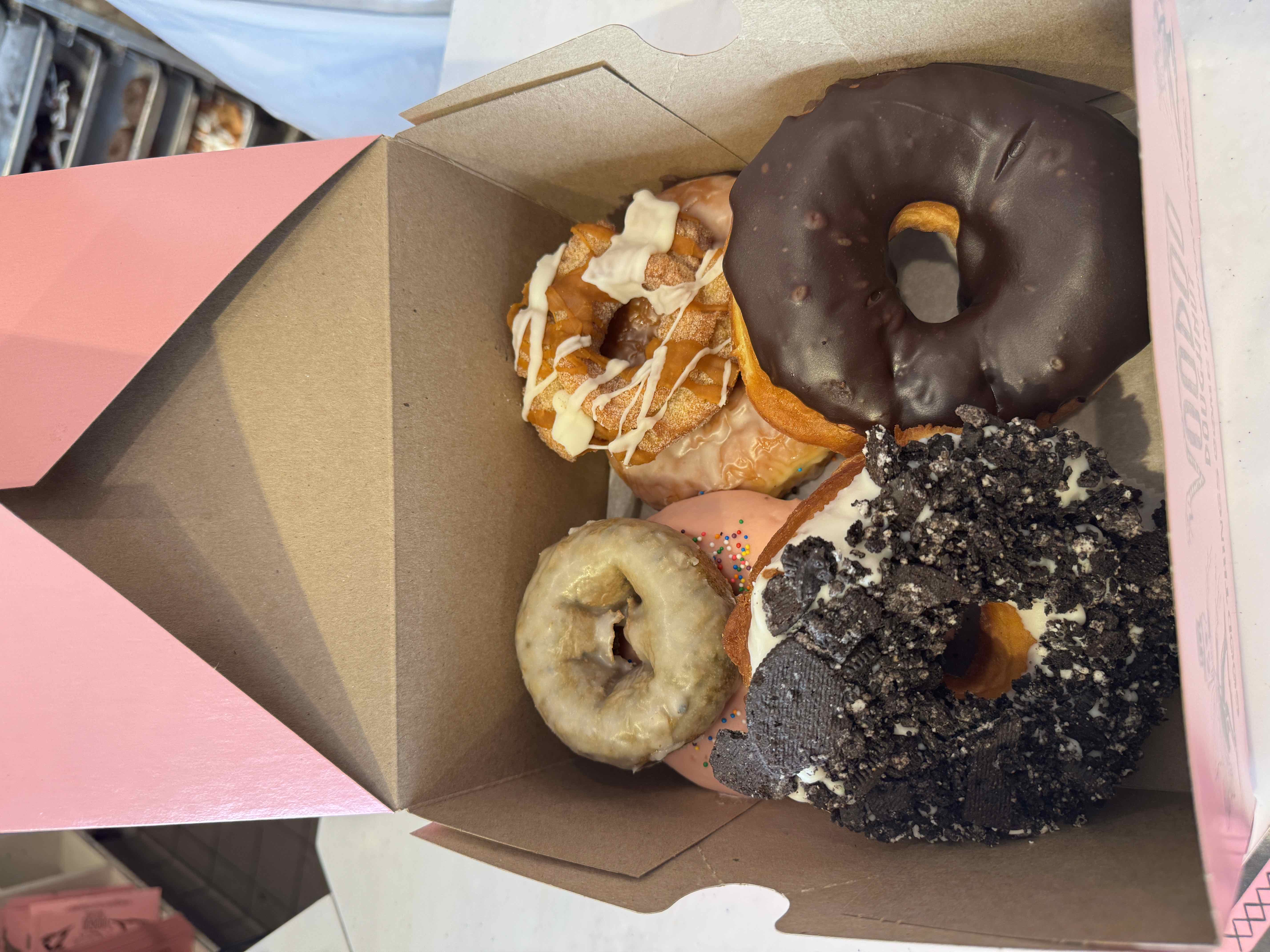 Image of a box of assorted donuts, including Oreo, chocolate glazed, and a pink sprinkle donut.