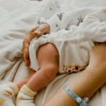 Close-up image of a newborn baby wearing socks, cradled by a person with a hospital wristband, on white bedding showing The 5 5 5 Postpartum Rule.