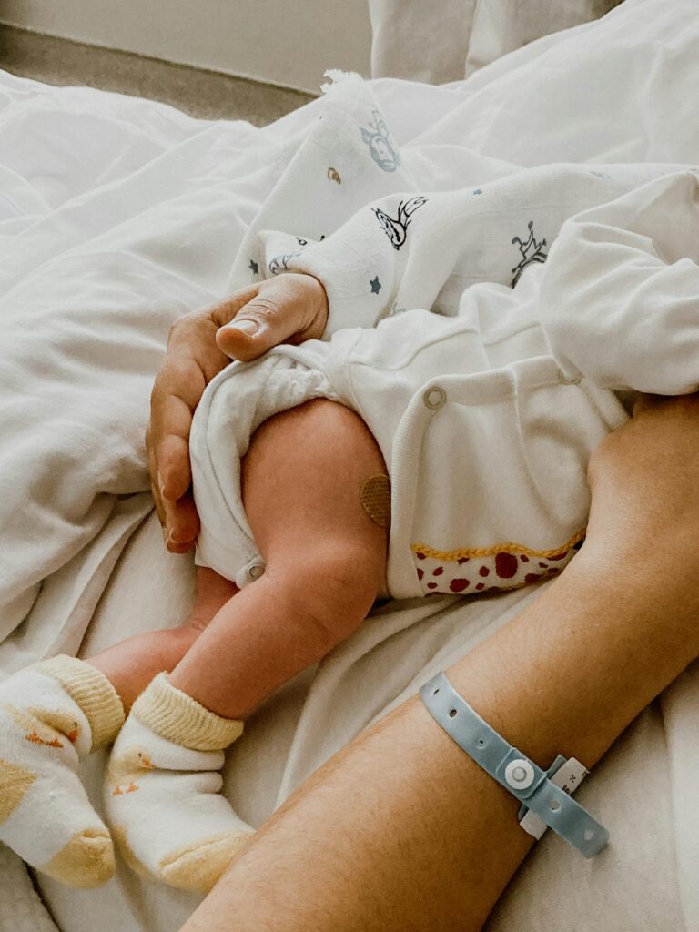 Close-up image of a newborn baby wearing socks, cradled by a person with a hospital wristband, on white bedding showing The 5 5 5 Postpartum Rule.