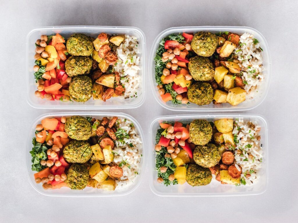 Image of four meal prep containers are filled with a colorful combination of falafel, roasted vegetables, white rice, and fresh chopped salad with chickpeas. The meals are neatly arranged on a light gray background showing Dinner ideas for tired moms.