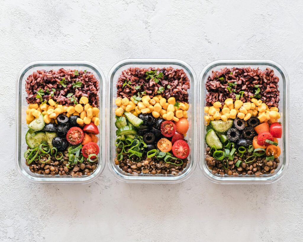 Image of three rectangular glass meal prep containers arranged side by side, each containing neatly layered ingredients: purple rice, yellow corn, black olives, sliced cucumbers, cherry tomatoes, green onions, and lentils, garnished with herbs.