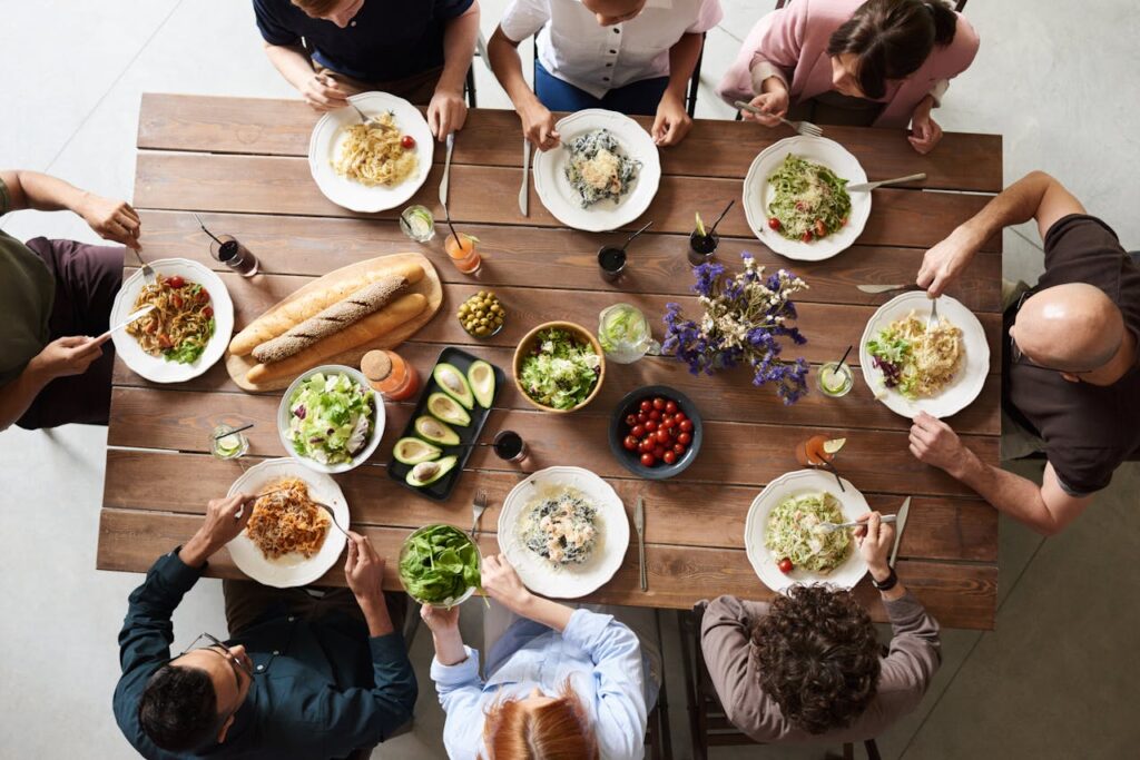 Image of a group of people seated around a wooden dining table sharing a meal, with plates of pasta, fresh salads, bread, avocados, olives, and cherry tomatoes, accompanied by drinks and a floral centerpiece, showing Meal Planning For Working Moms.