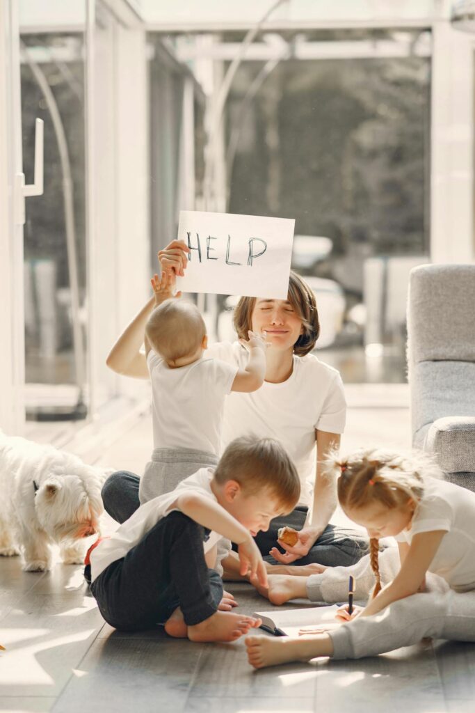 Image of a mother sitting on the floor with kids and a dog holds a HELP sign, surrounded by chaos, representing A Day in the Life of a Mom.